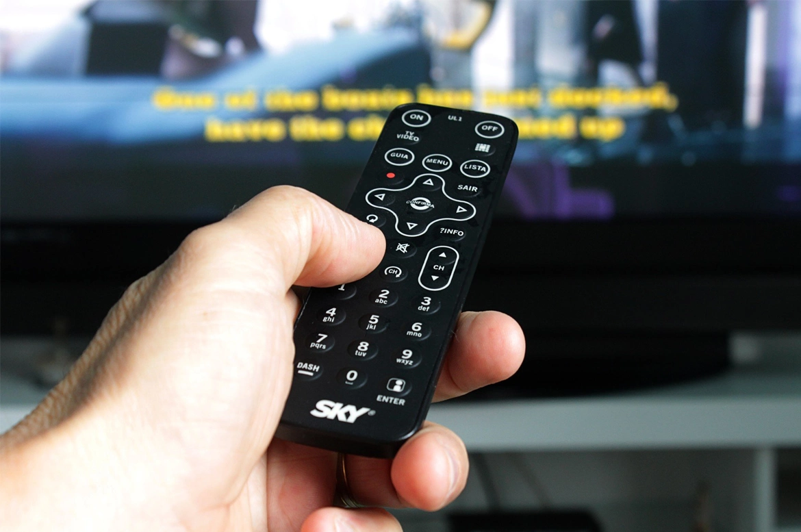 A hand holds a remote control pointed towards a television, while subtitles appear on the screen in the background.